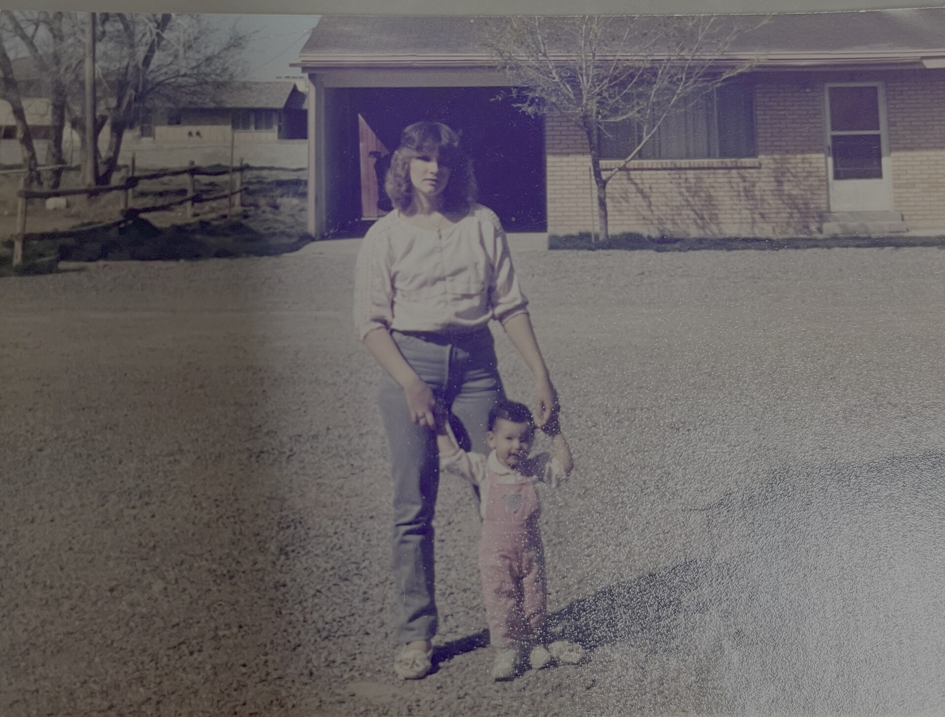 Jen as a toddler in overalls, holding her mom's hands on a gravel driveway in front of a brick house.