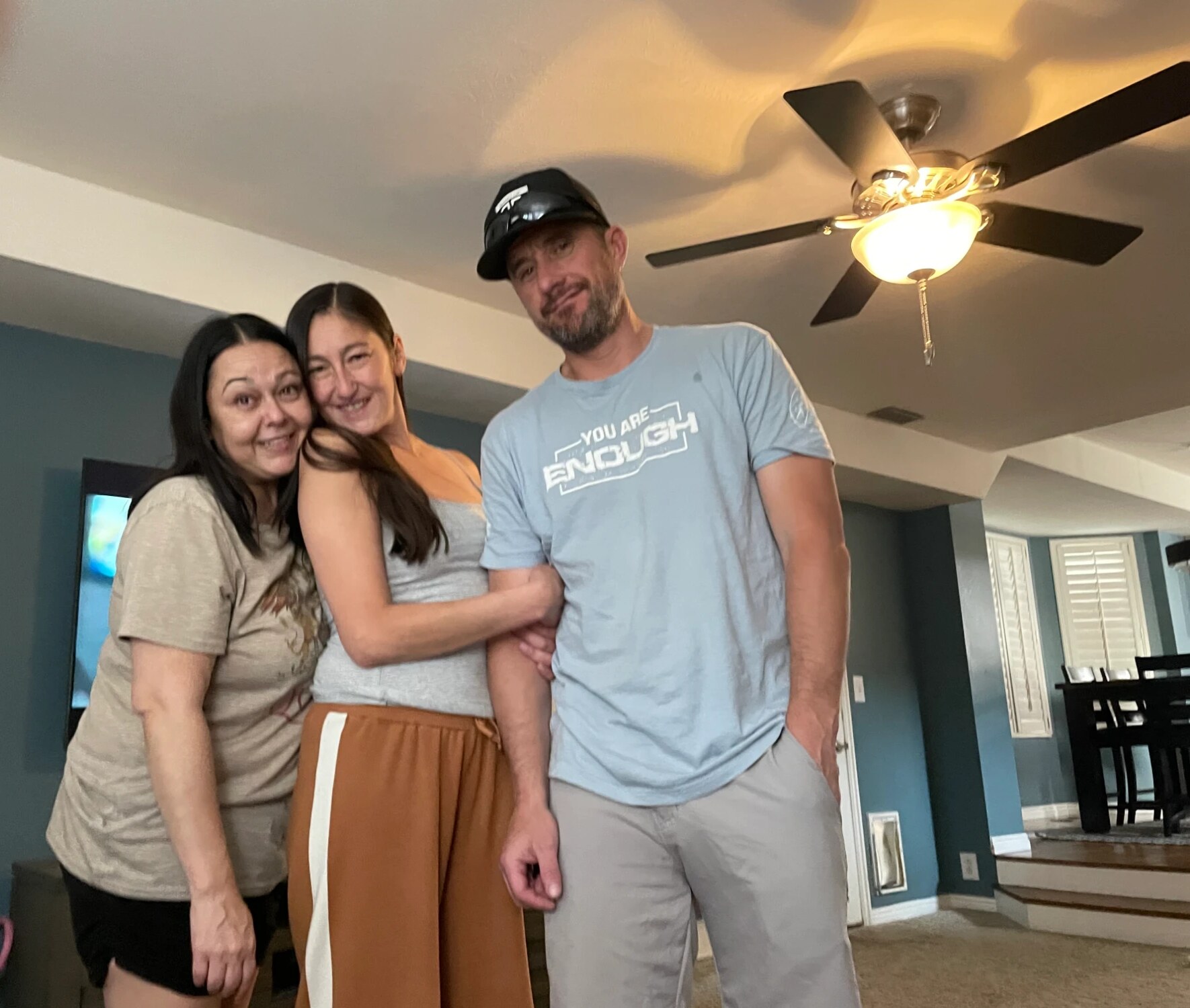 Jen between her sister and her brother, hugging his arm, the three of them in a living room.